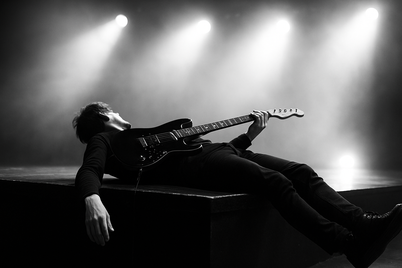 the guitar is lying across the body, one hand holds the neck of the guitar and the other hand is hanging off the side of the stage. the head of the person is looking away.